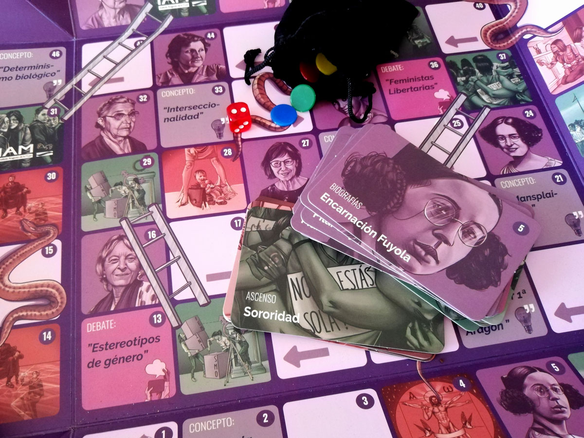 En esta imagen se observa el tablero del juego con la diversidad de sus casillas las escaleras y las serpientes. Encima del tablero hay situado un mazo de cartas de biografías y conceptos a modo de muestra de los objetos en los que se compone el juego