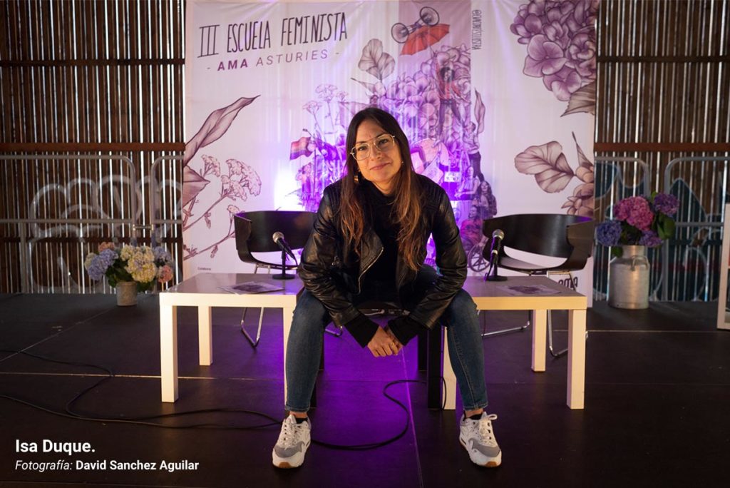 Isa Duque, retratada sobre el escenario de la III Escuela Feminista AMA Asturies, con la gráfica de fondo de Eva Cortés Ilustra